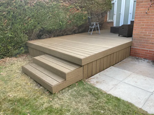 Raised composite deck and steps with light-brown wood-grain finish, installed against a brick property.